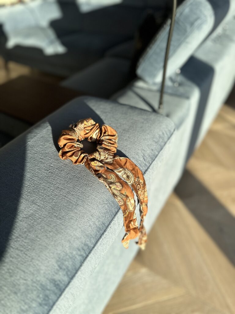 Boho Cacao scrunchie s mašlí položená na opěrce světle modré pohovky, ozářená sluncem.