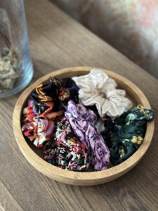 Miska s různými barevnými scrunchies z viskózy a saténu položená na dřevěném stole, mix vzorů a přírodních materiálů.