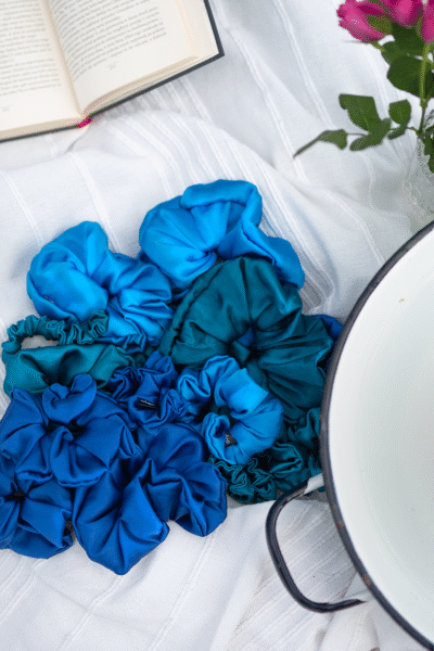 Modré hedvábné scrunchies Caloo v různých odstínech položené na bílé látce vedle knihy, květin a smaltované misky, foceno v přirozeném světle.