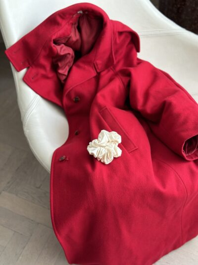 Krémová saténová scrunchie Caloo Satin Pearl položená na sytě červeném kabátu umístěném na bílé designové židli, elegantní styling pro produktovou prezentaci.