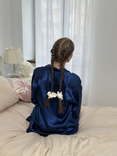 Dívka sedící na posteli v modrém hedvábném županu, s copánky sepnutými dvěma béžovými saténovými scrunchies Caloo Satin Pearl, foceno v jemném denním světle.