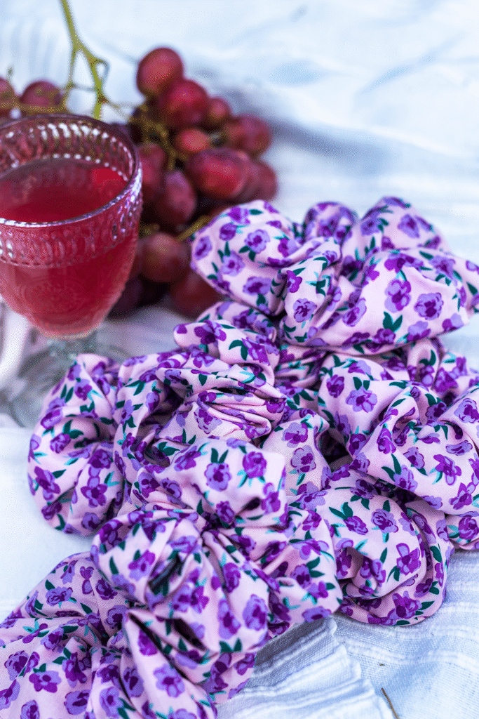 Gumičky Purple Blossom z viskózy s květinovým potiskem, naaranžované na látce vedle sklenice vína a hroznů