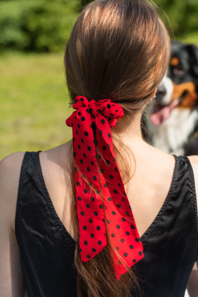 Gumička Polka Dot z viskózy s dlouhou červenou mašlí s černými puntíky ve vlasech – nízký culík, pozadí příroda, v pozadí pes